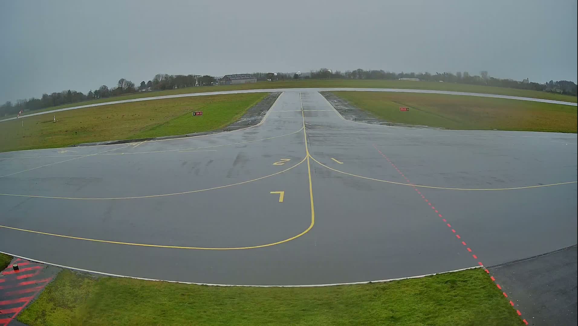 A wet, gray airport tarmac with yellow and red markings stretches toward a runway on an overcast day.