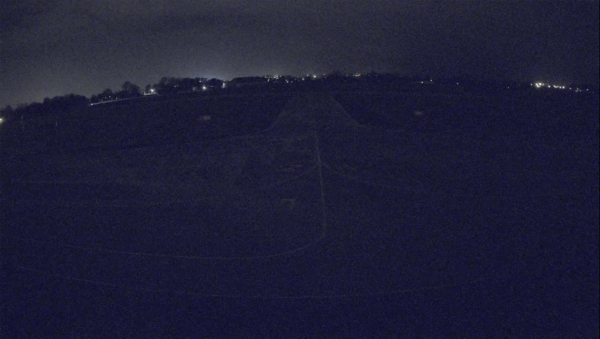 A dark, nighttime view of a flat, open expanse of land with a faint road or path leading towards a distant line of dimly lit buildings under a clear, dark sky.