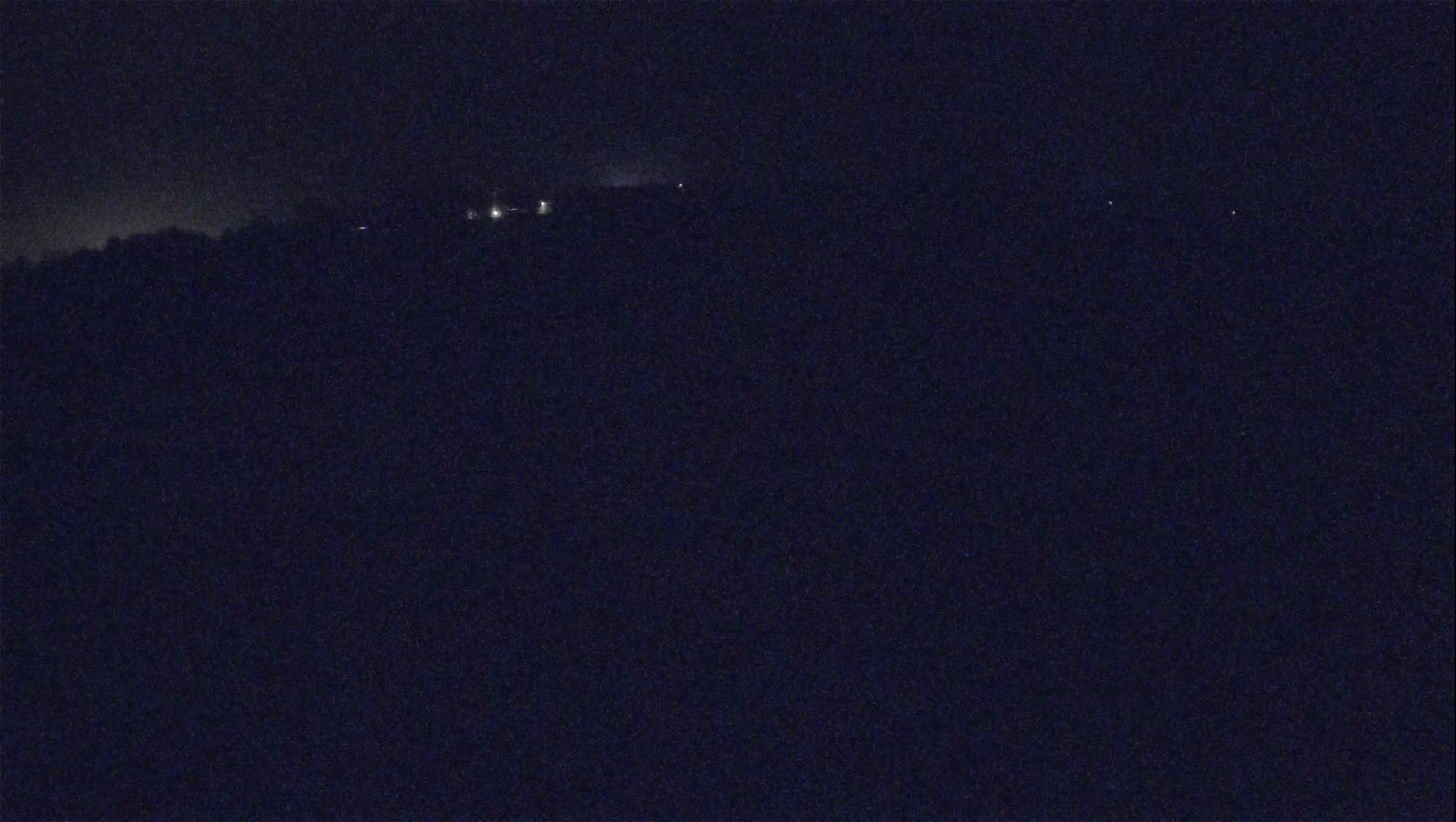 A dark, nighttime view shows a distant line of faint lights atop a dark hill or ridge under a clear, starless sky.