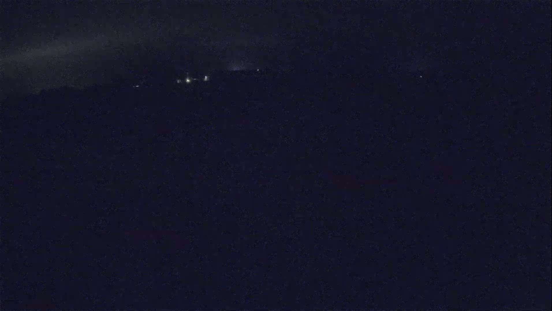 A dark, nighttime landscape shows a few faint lights in the distance under a cloudy sky.