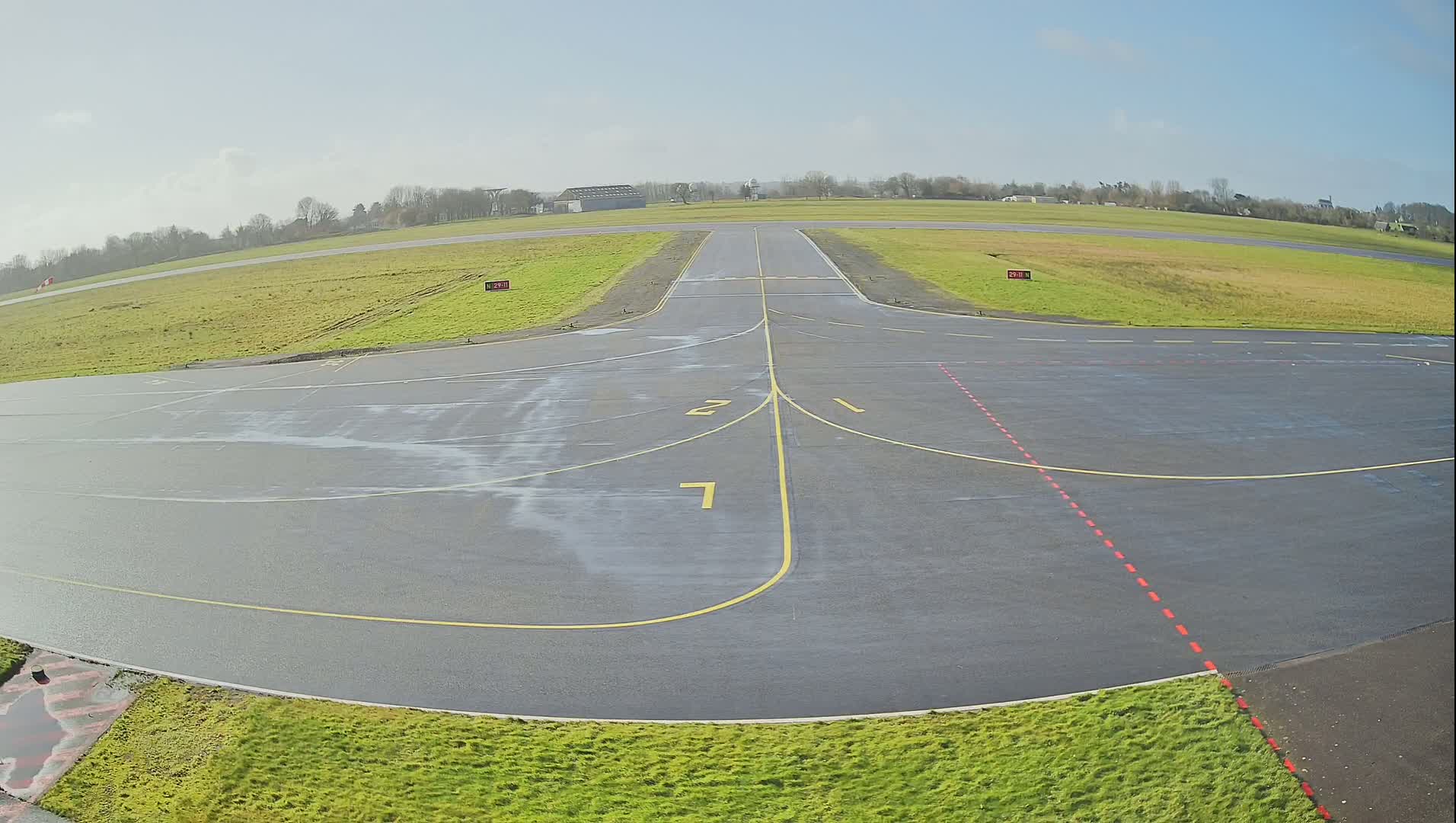 A wet airport tarmac with yellow and red markings is visible, extending to a grassy field under a bright, mostly clear sky.
