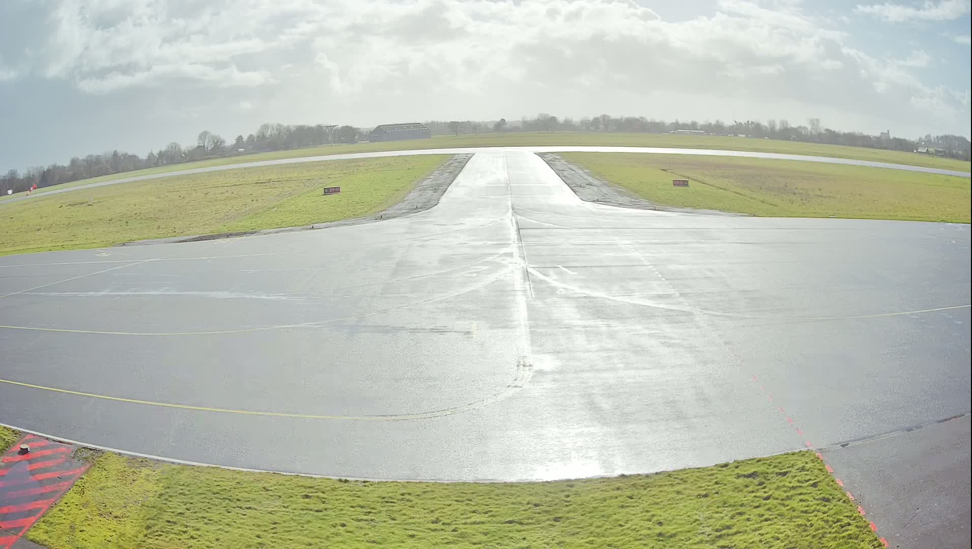 A wet, paved airfield runway extends to the horizon under a partly cloudy sky.