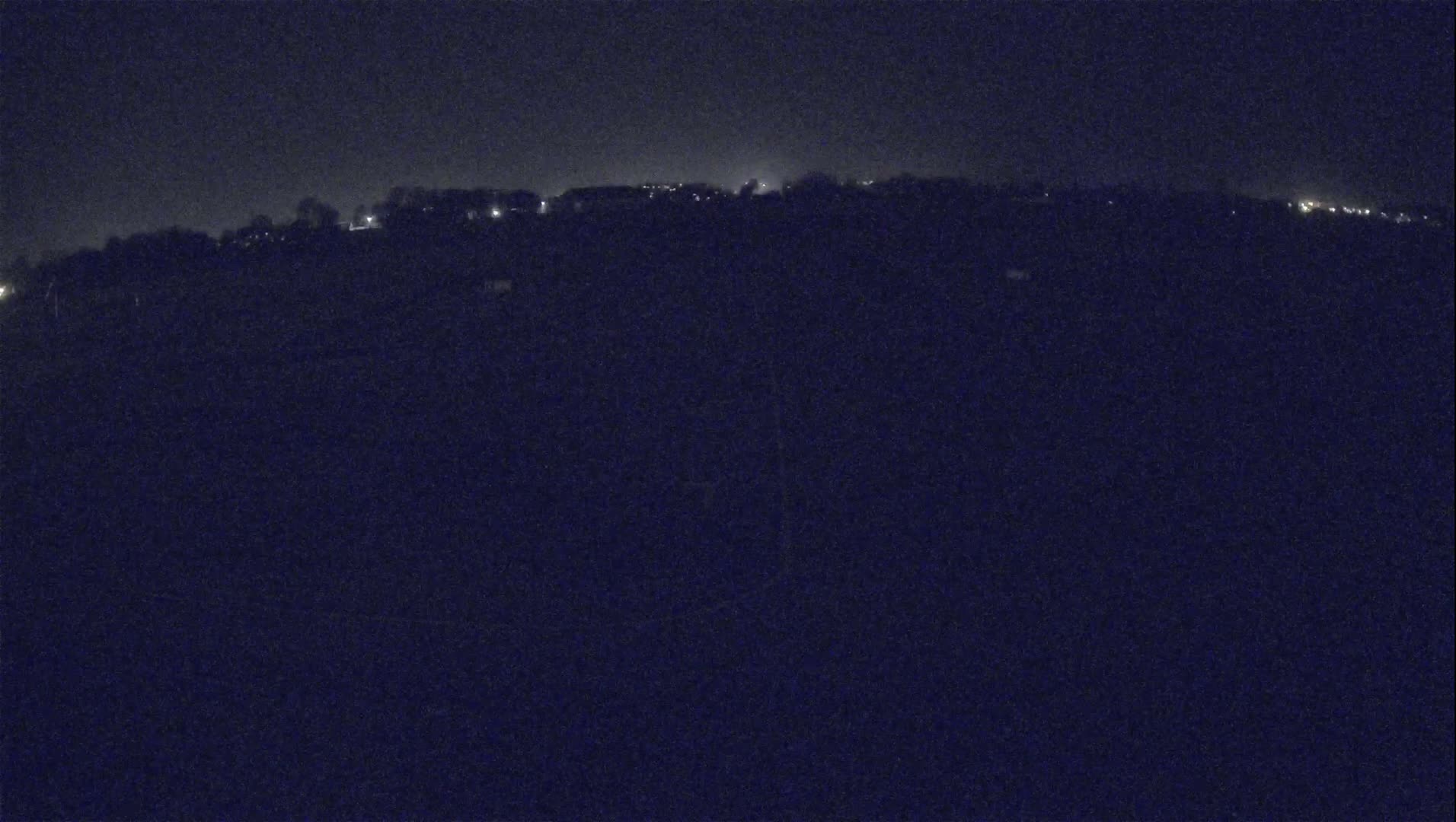 A dark, nighttime landscape shows a distant line of dimly lit buildings on a hill under a clear, dark sky.