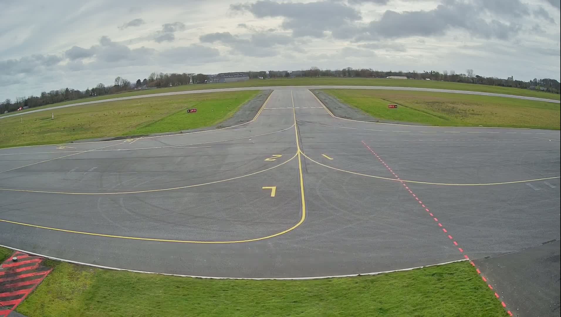 An empty airport tarmac under a cloudy sky shows a runway intersecting with taxiways marked by yellow lines.