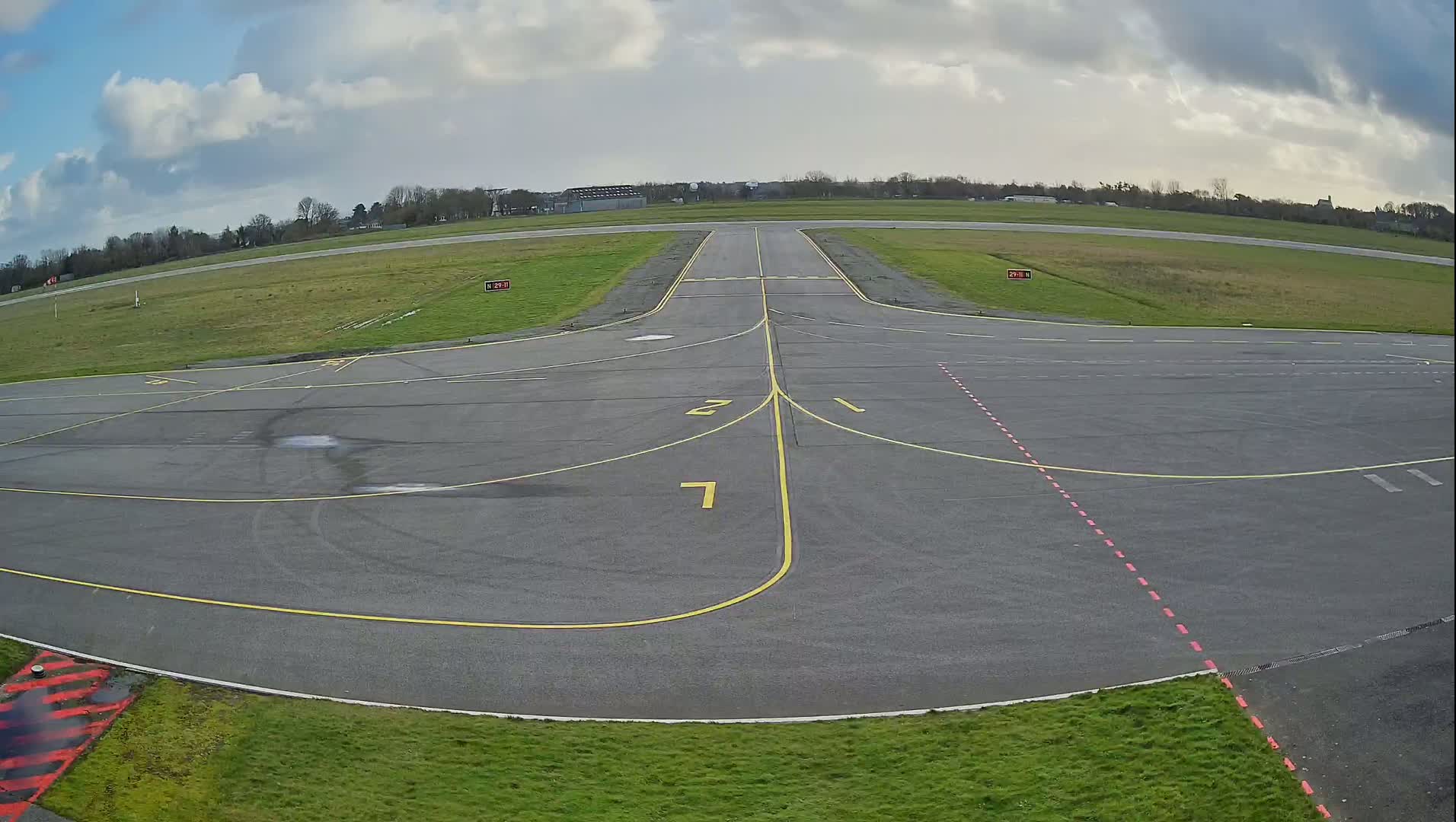 A mostly cloudy day at an airport shows an empty tarmac with yellow and red markings, and a runway extending into the distance.