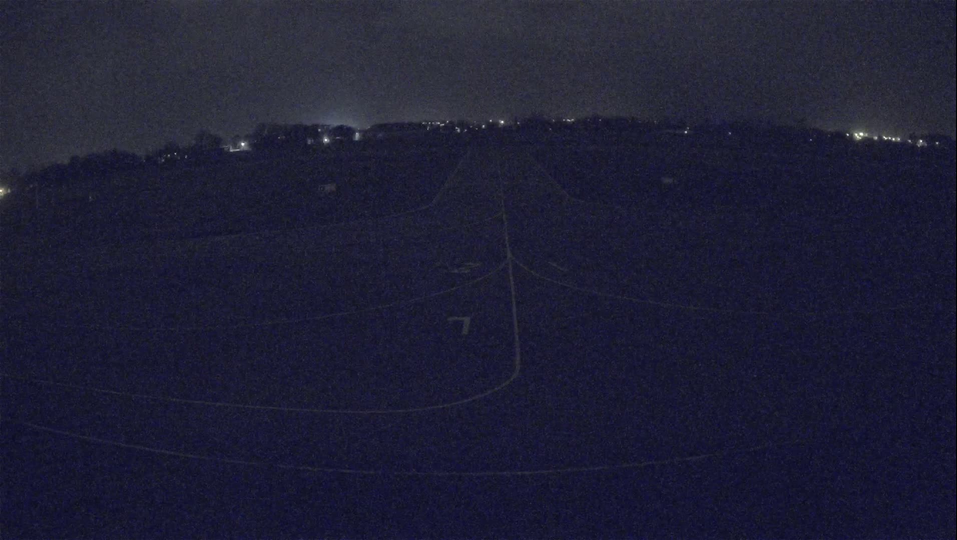 A dark, nighttime view of a field with faint white lines marking its sections, sparsely lit by distant lights under a cloudy sky.