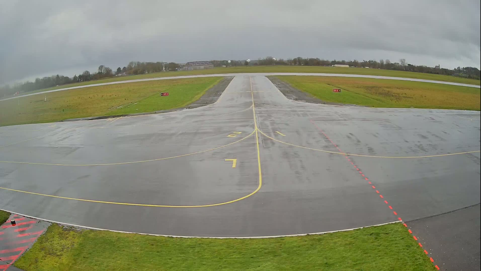 A wet, gray airport tarmac with yellow runway markings is shown under an overcast sky.