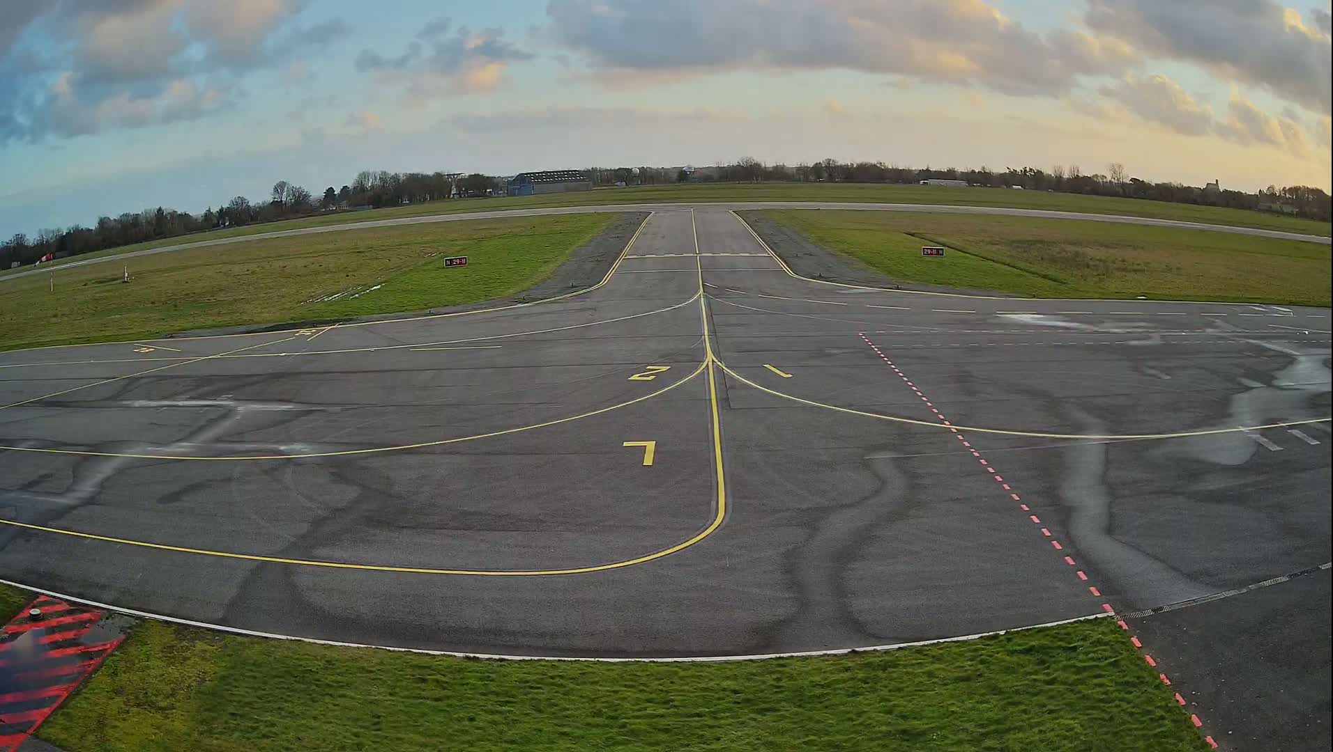 An airfield's tarmac, marked with yellow lines and numbers, stretches towards a grassy horizon under a partly cloudy sky.