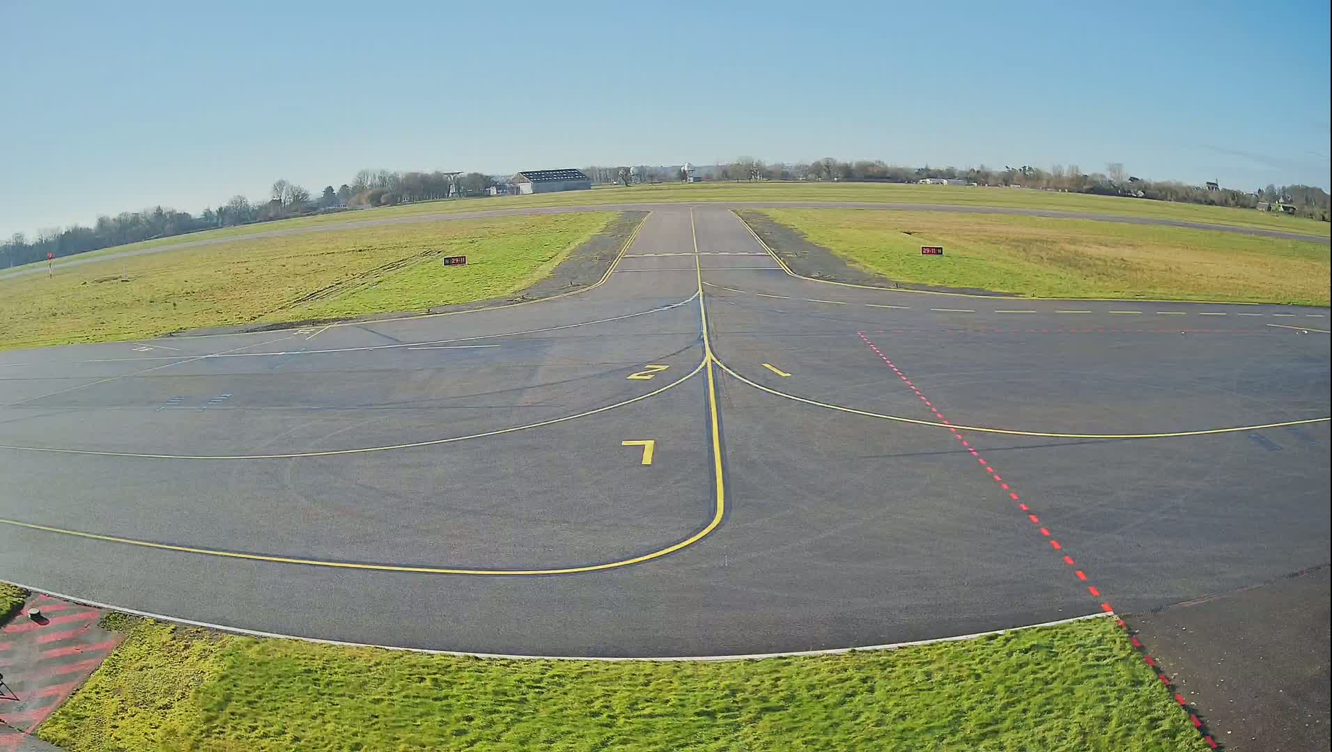 A tarmac airport runway and taxiway area is shown on a sunny day, surrounded by short, green grass.
