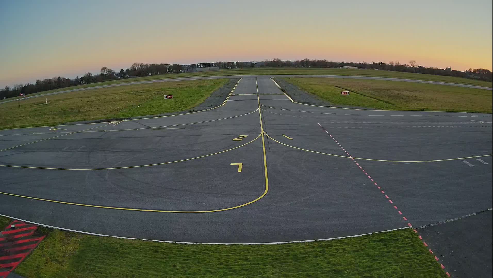 An empty airport tarmac under a pale sunset sky shows runways and taxiways marked with yellow lines and red dashed lines.