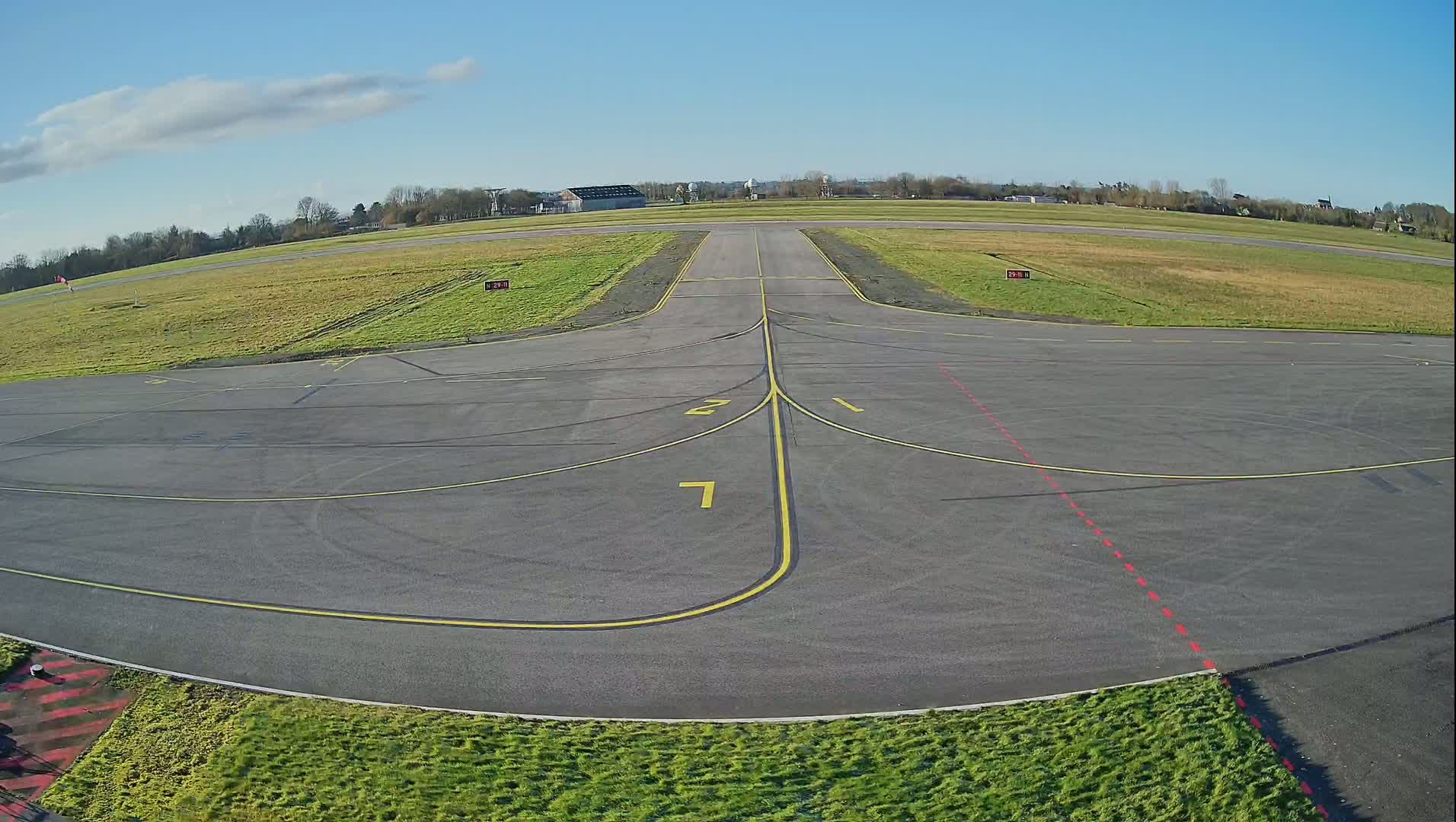 A tarmac airplane taxiway with yellow and red markings is surrounded by short green grass under a mostly clear blue sky.