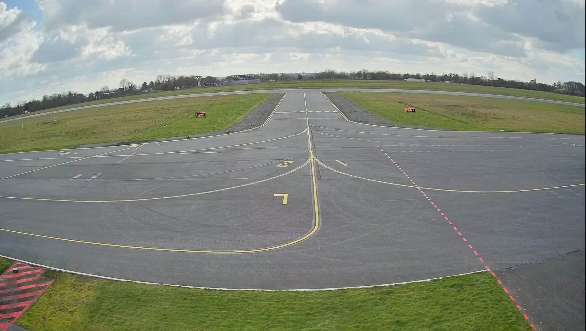 A mostly cloudy day reveals an airport tarmac with various yellow and red markings, a runway extending into the distance, and green grass surrounding the paved area.