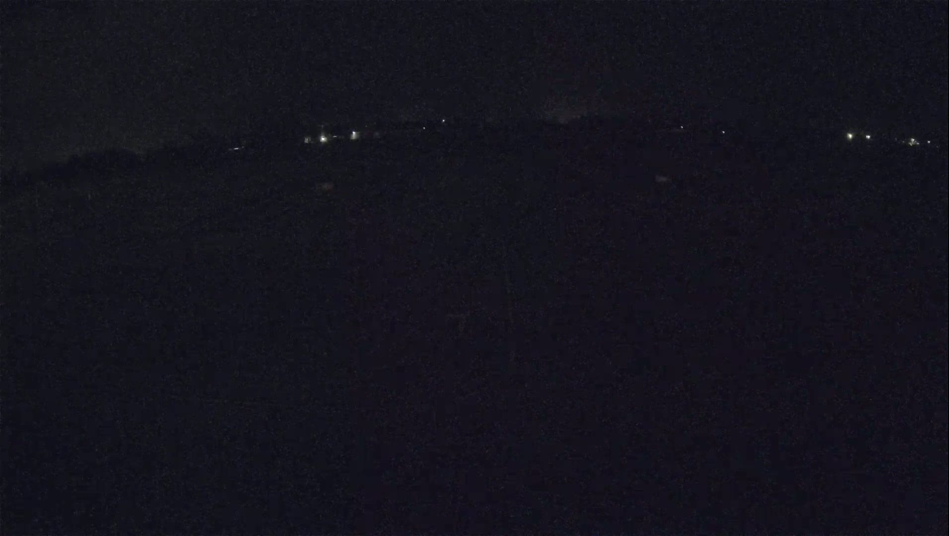 A dark, nighttime landscape shows a distant line of faint lights under a mostly clear sky.