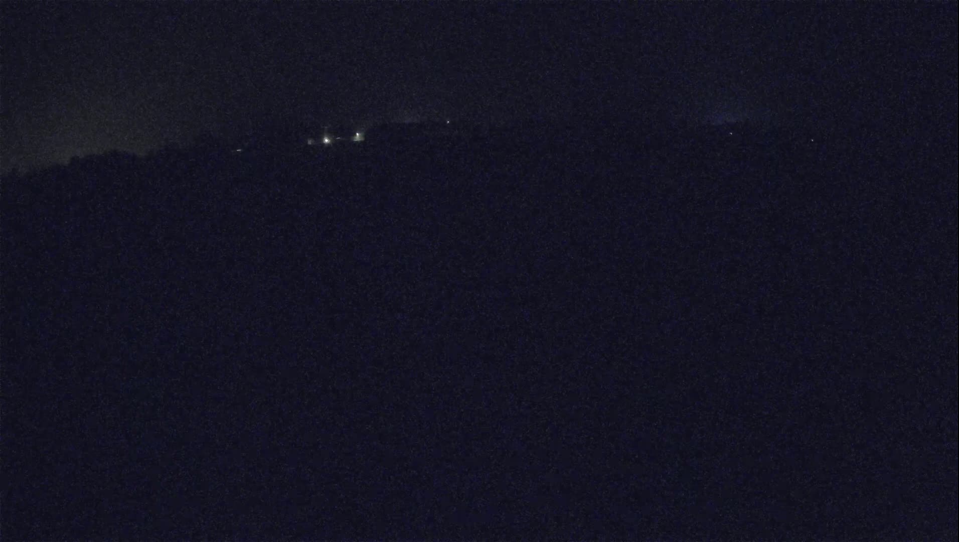 A dark, nighttime landscape shows a distant line of faint lights on a hill under a dark, possibly cloudy sky.