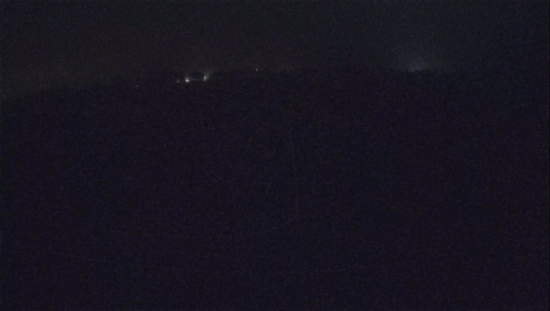 A nighttime long shot shows a dark landscape with a few faint lights in the distance under a dark sky.