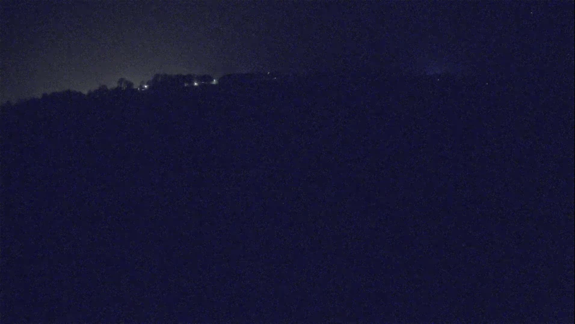 A dark, nighttime view of a distant hilltop with a few faint lights visible against a mostly clear, black sky.
