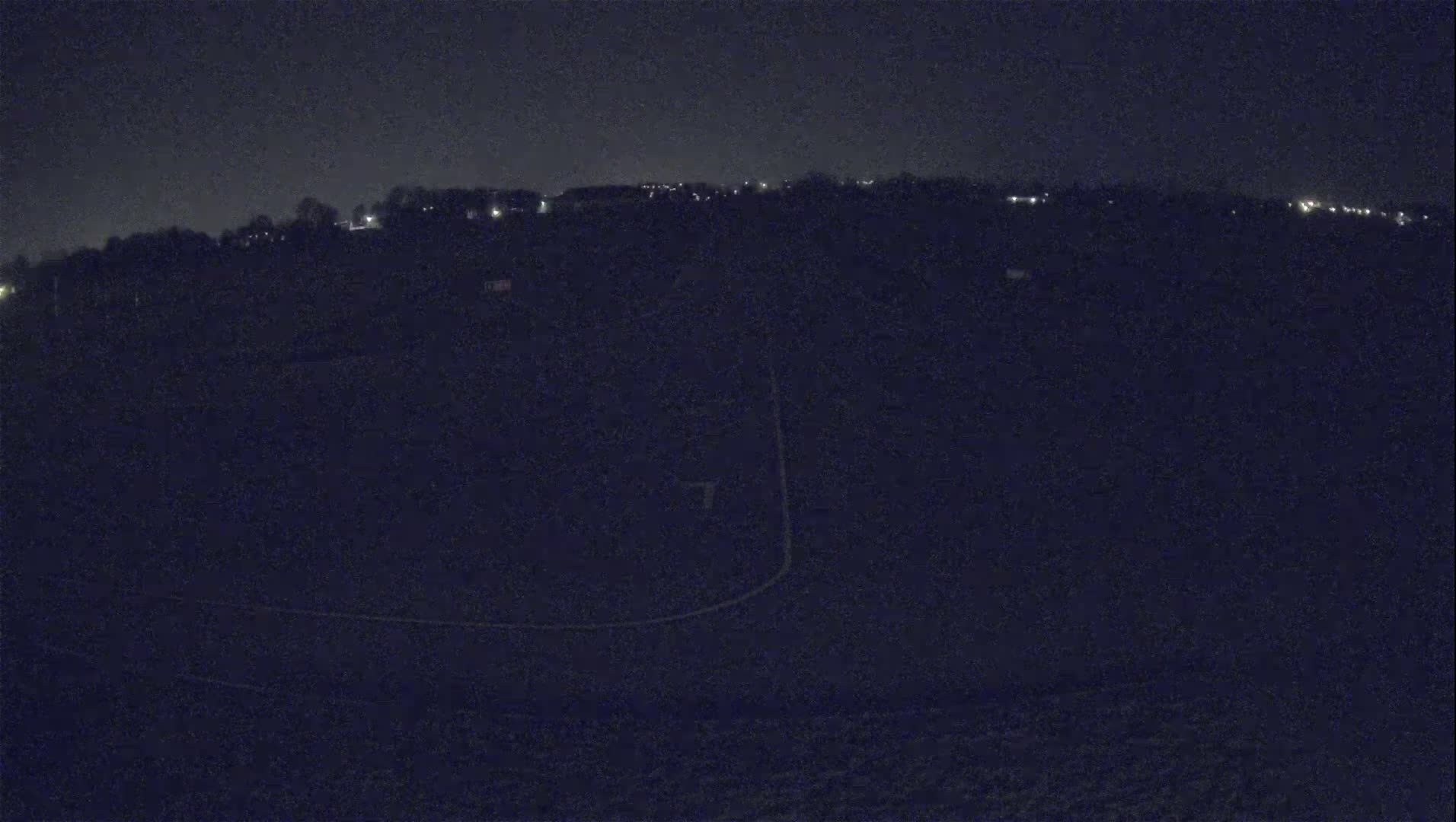 A dark, nighttime view of a hilly landscape with sparsely spaced lights in the distance and faintly visible outlines of what appear to be roads or paths in the foreground.