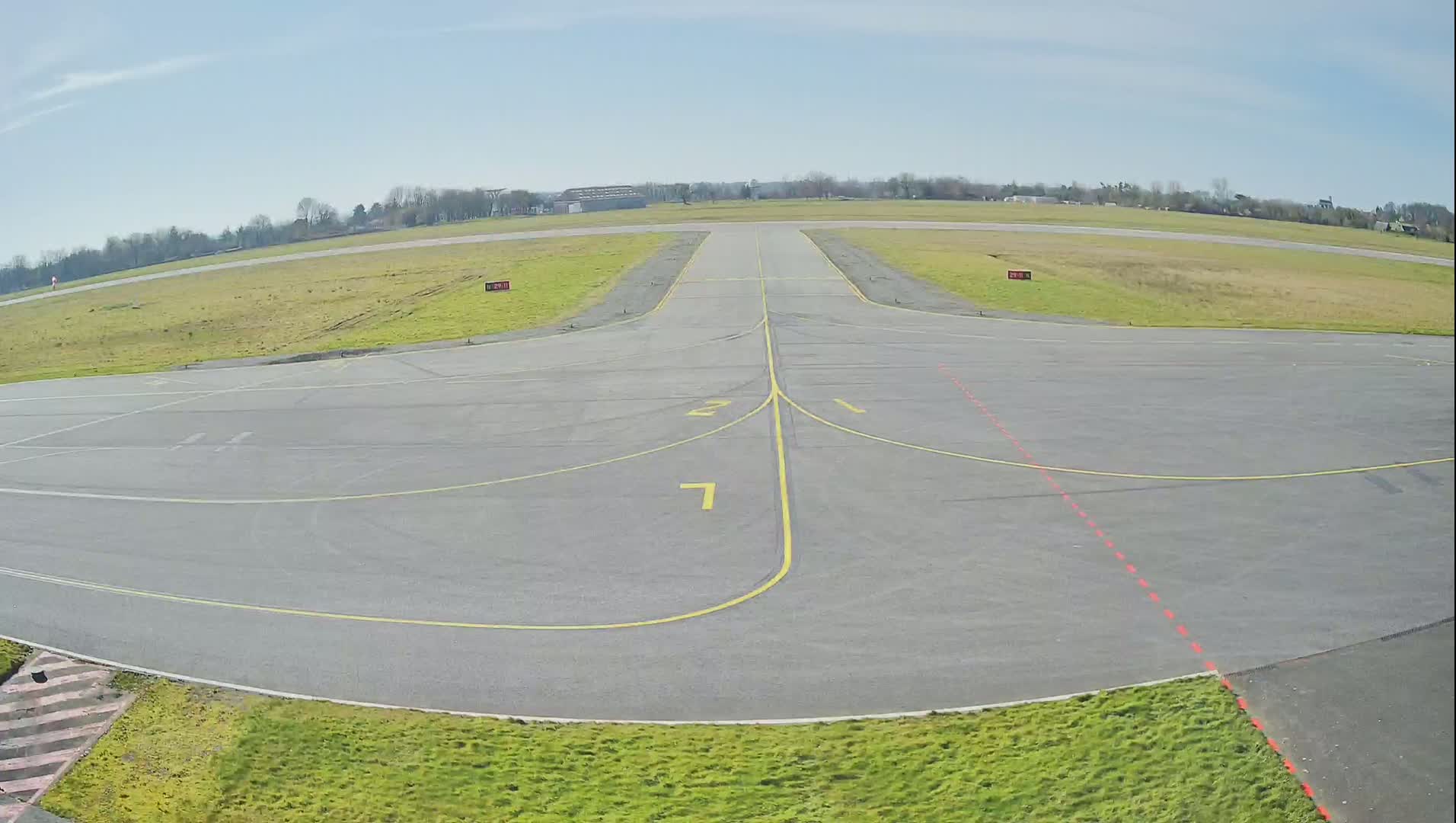 An airfield's tarmac, marked with yellow lines and numbers, stretches toward a grassy runway under a clear, bright sky.