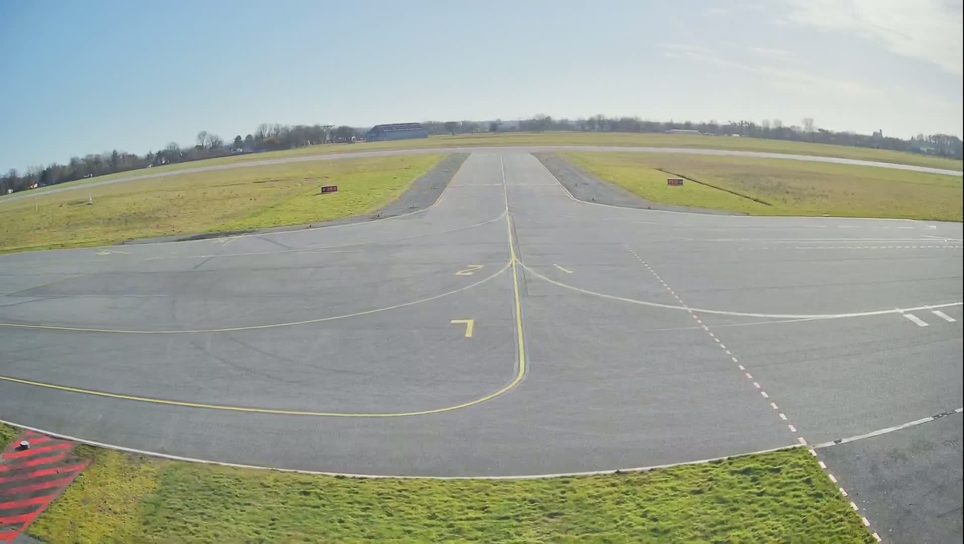 A tarmac airplane runway and taxiway area is visible on a sunny day, with short grass bordering the paved surfaces and a treeline in the background.