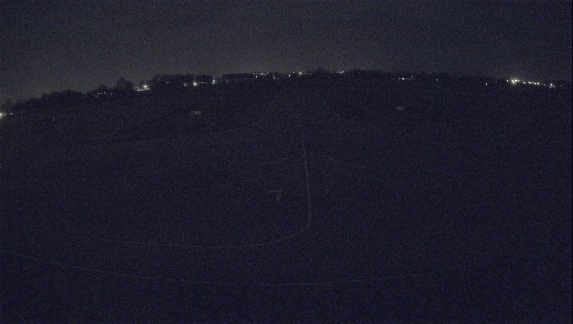 A nighttime, low-light view of a dark field with faint outlines of paths and a distant, sparsely lit horizon.