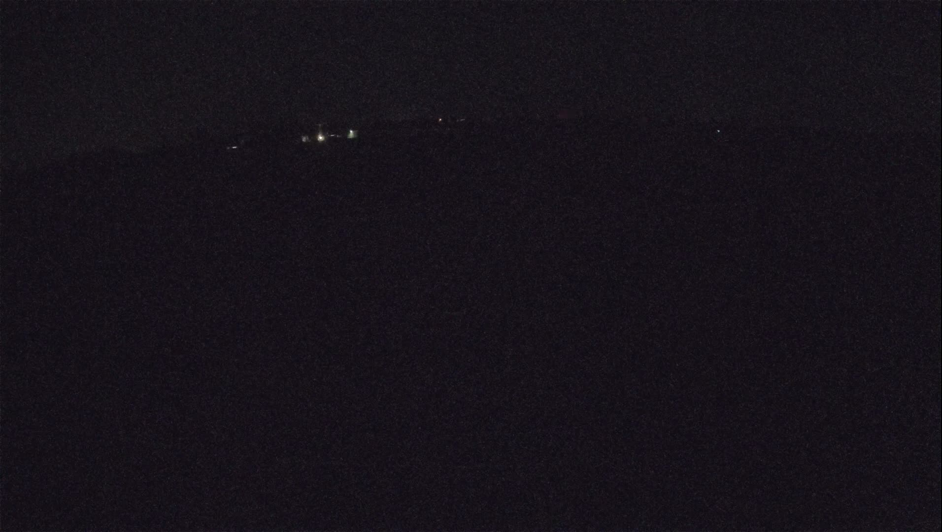 A nighttime view shows a dark landscape with a few faint lights in the distance under a dark sky.