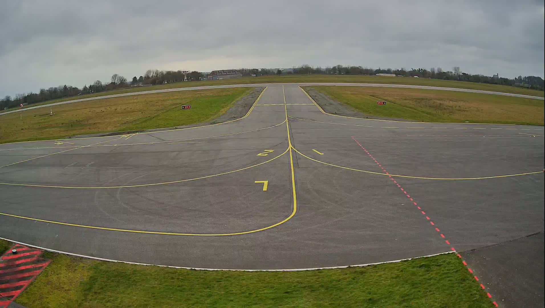 An overcast sky hangs over a small airport's tarmac, which shows taxiways marked with yellow lines and numbers.