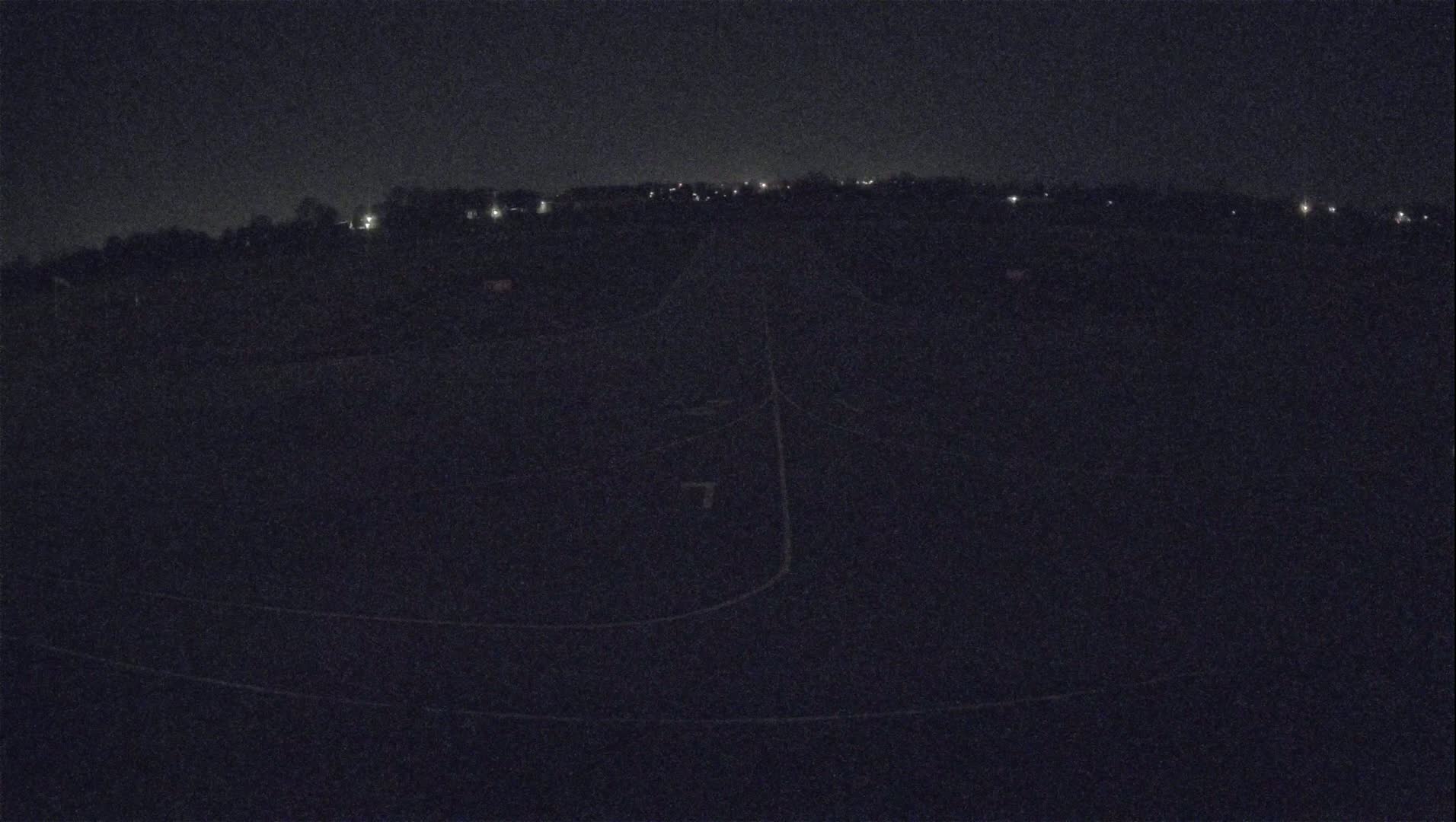 A dark, nighttime aerial view shows a paved surface with faint markings, possibly a road or runway, surrounded by a dark landscape with sparsely scattered distant lights.