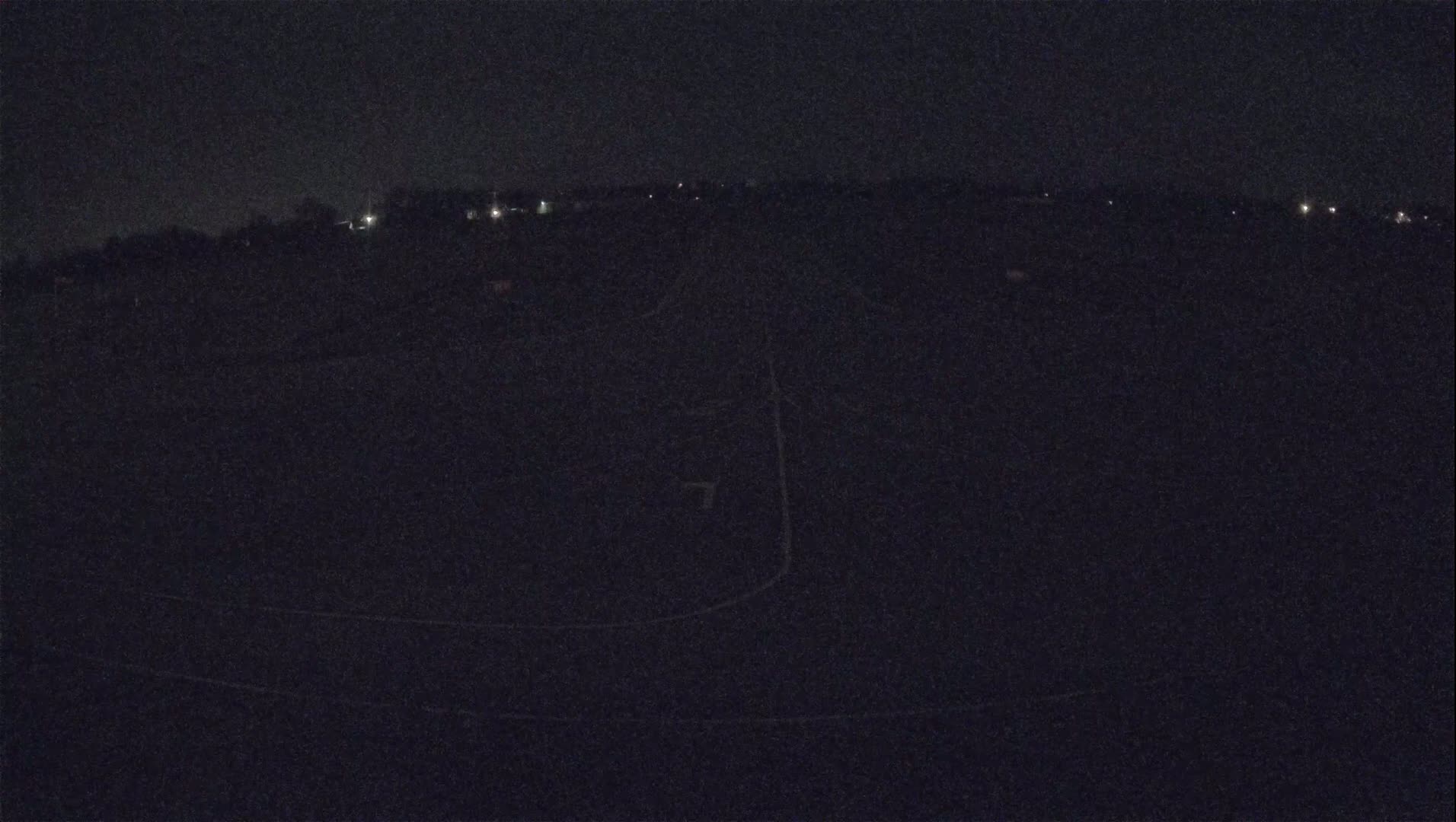 A dark, nighttime view of a road curving through a field with a few distant lights visible.