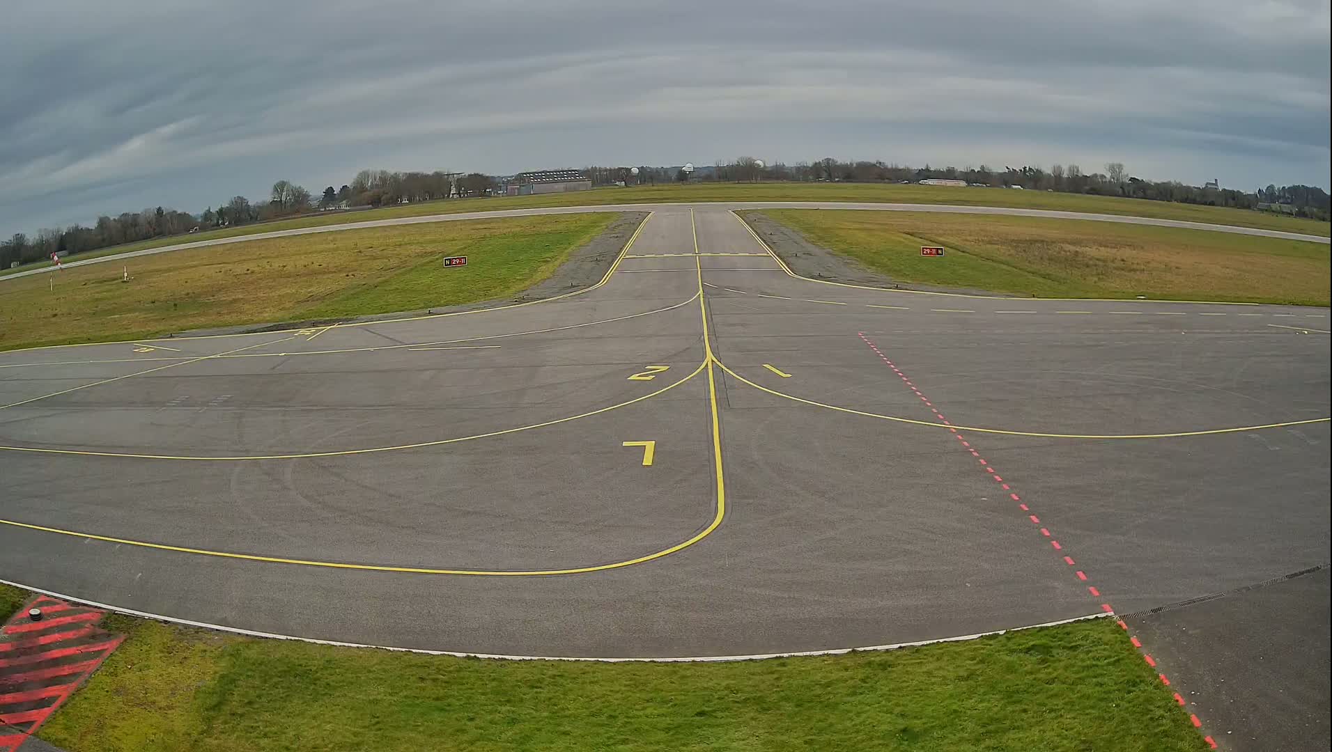 An airfield apron with yellow and red markings, under a cloudy sky, shows a runway extending into the distance.