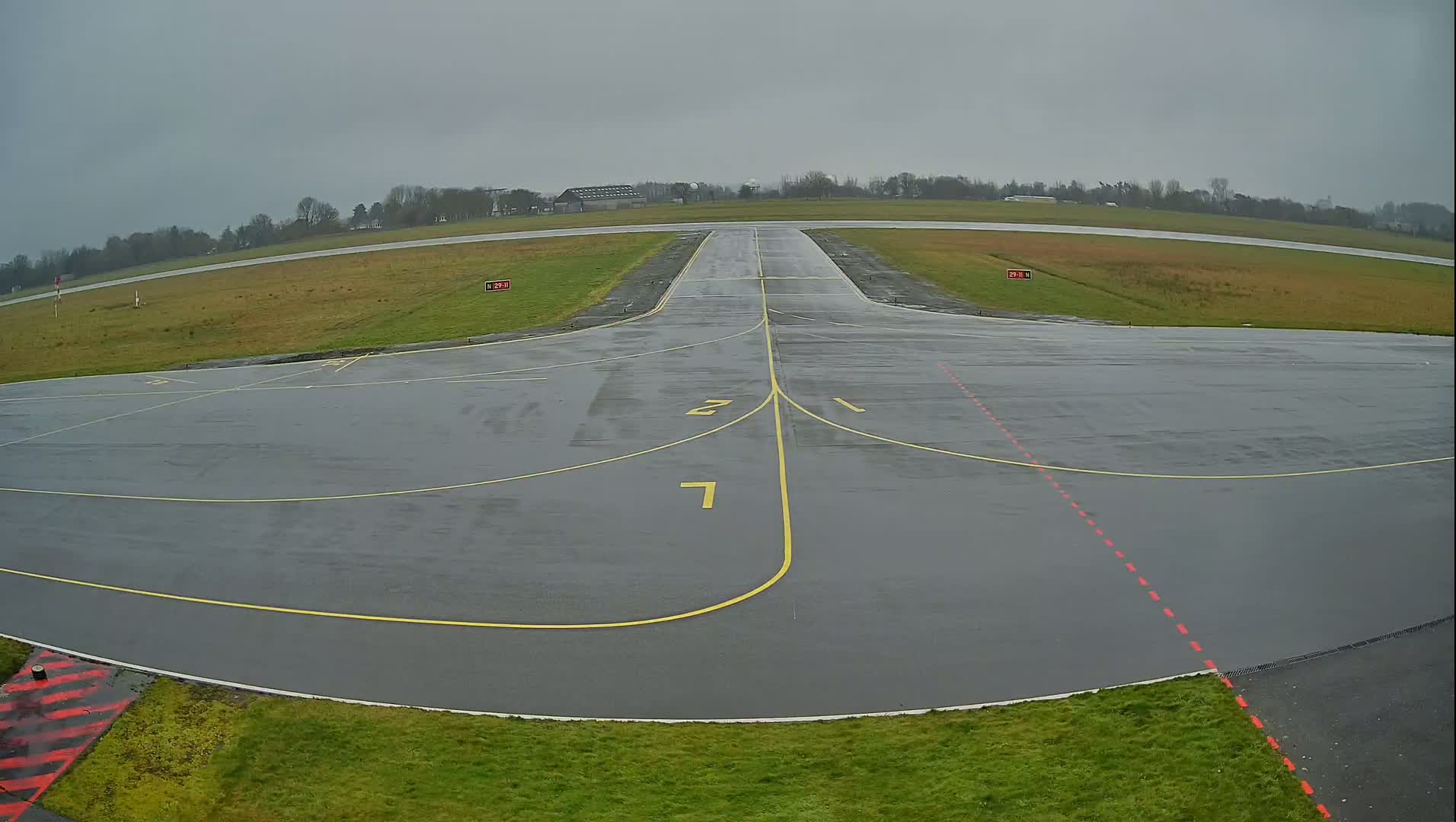 A wet, gray airport tarmac with yellow lines marking taxiways and runways, extending to a grassy field under an overcast sky.
