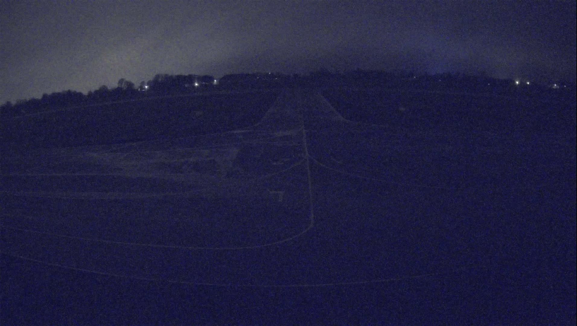 A dark, nighttime view of a field with a road or path cutting through it, faintly illuminated by distant lights.