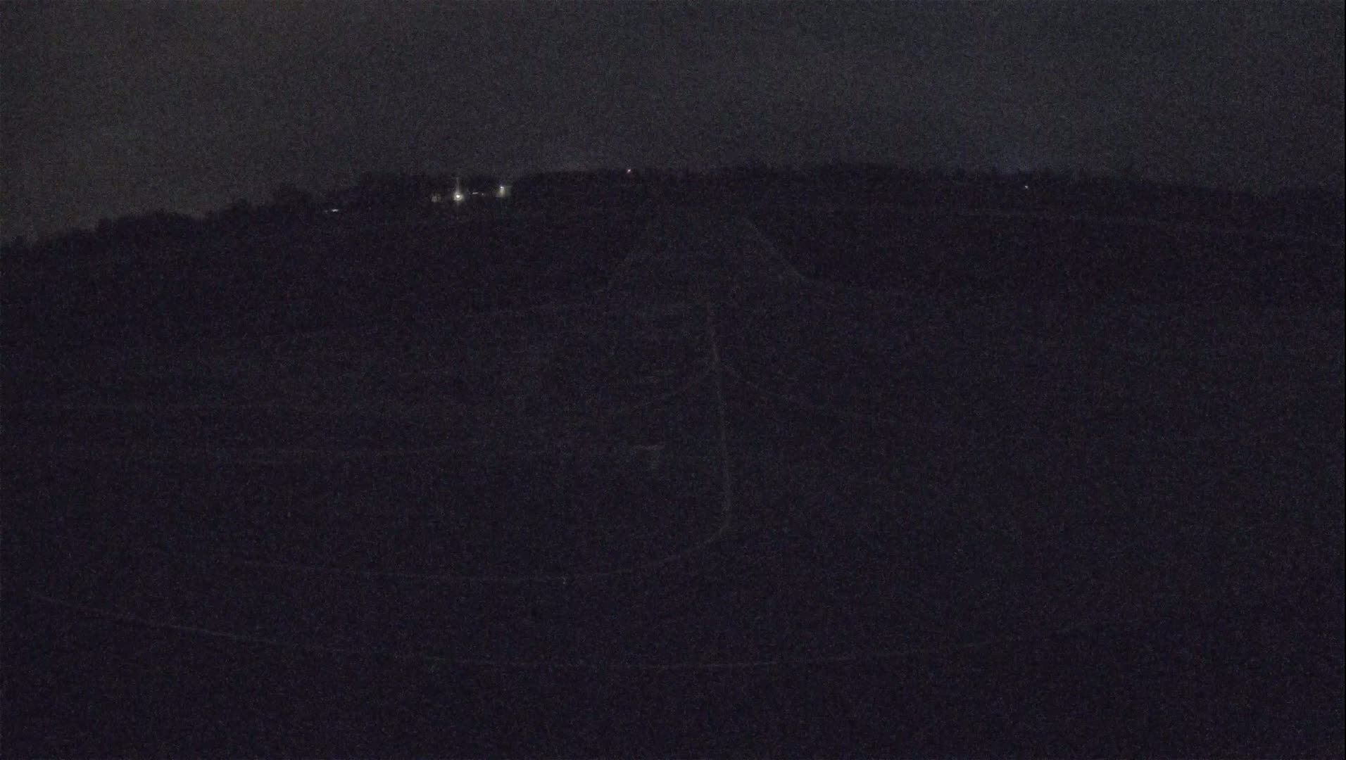 A dark, nighttime view of a mostly flat landscape with a few distant lights visible under a cloudy sky.