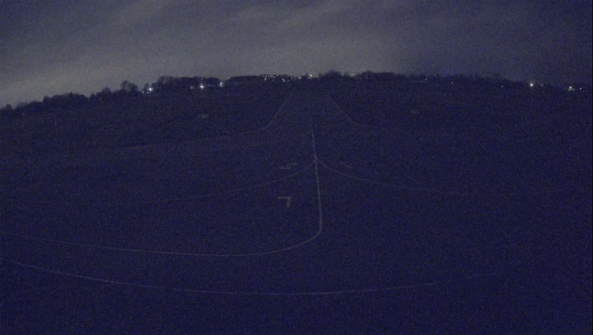 A dark, nighttime view of a paved runway or road with faint markings, surrounded by a treeline in the distance under a cloudy sky.