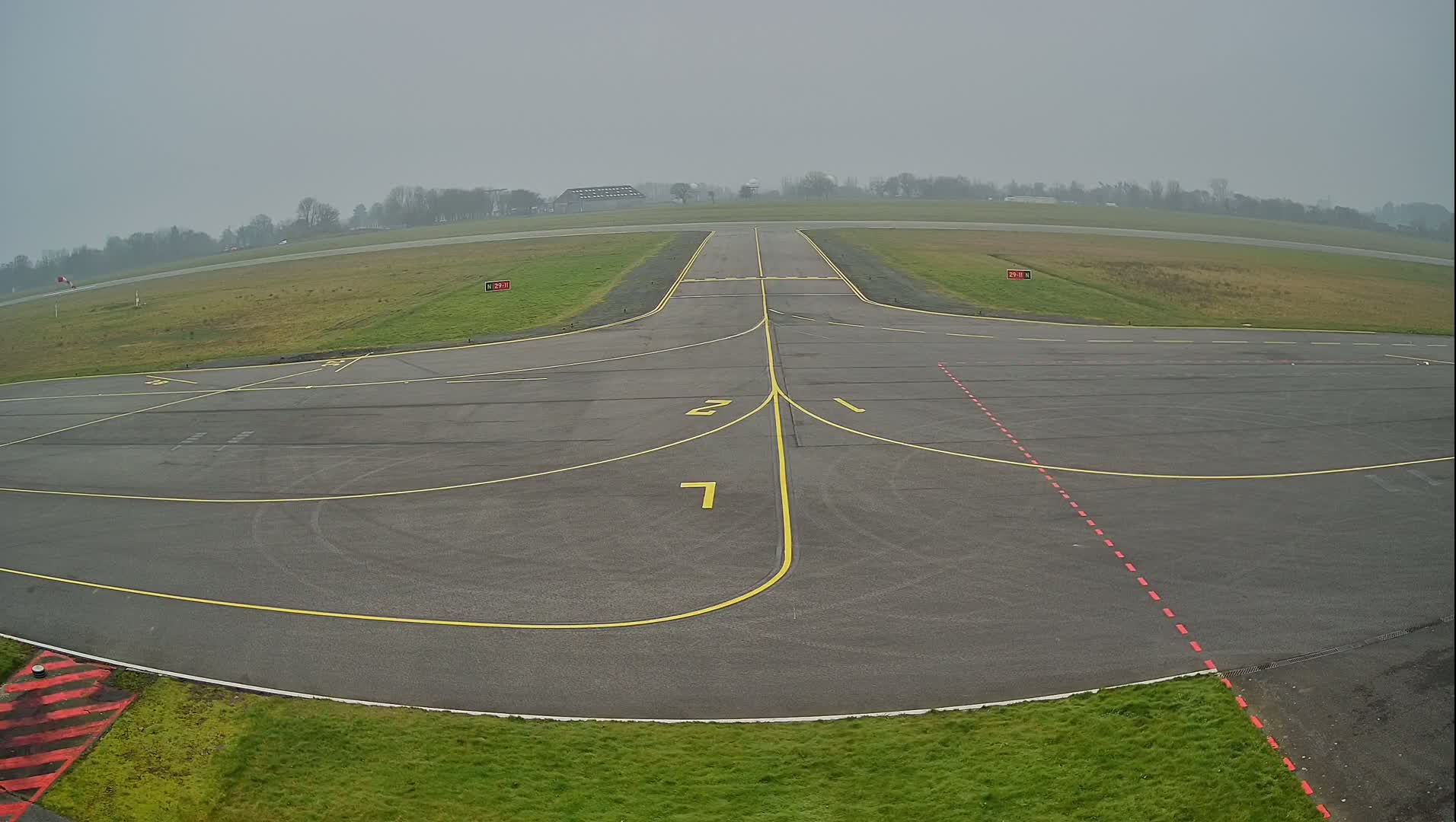 A foggy view of an airport runway and taxiway with yellow and red markings on the gray tarmac, surrounded by short green grass.