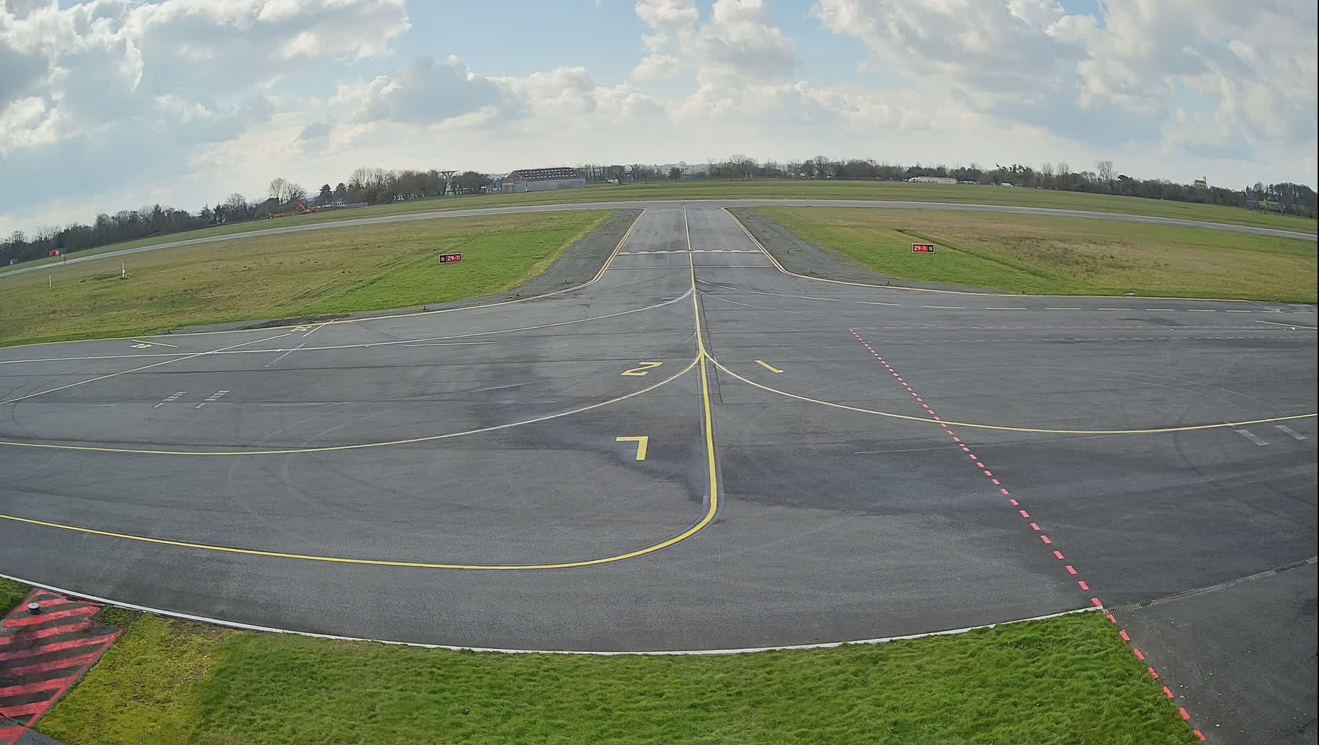 An airfield's tarmac apron with yellow and red markings, under a partly cloudy sky.