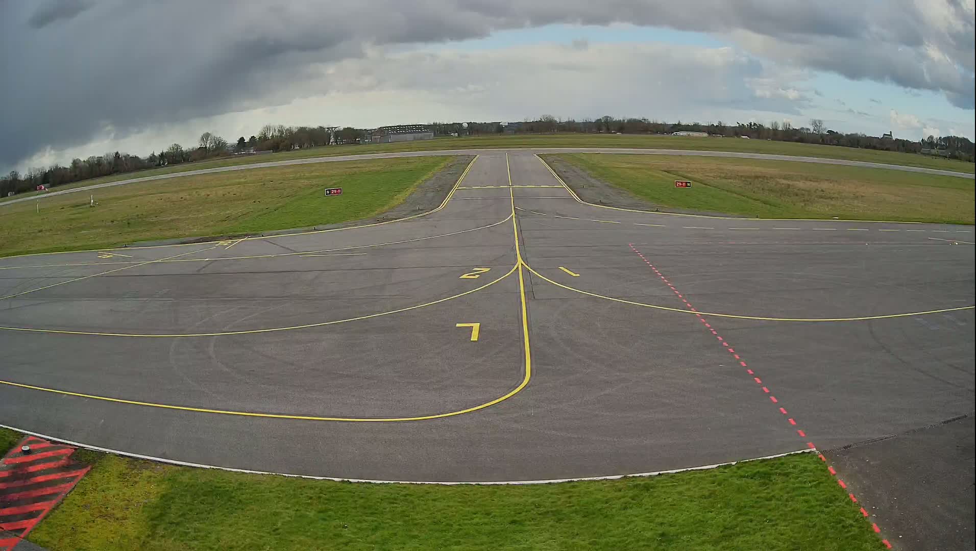An airfield taxiway with yellow and red markings is seen under a cloudy sky.