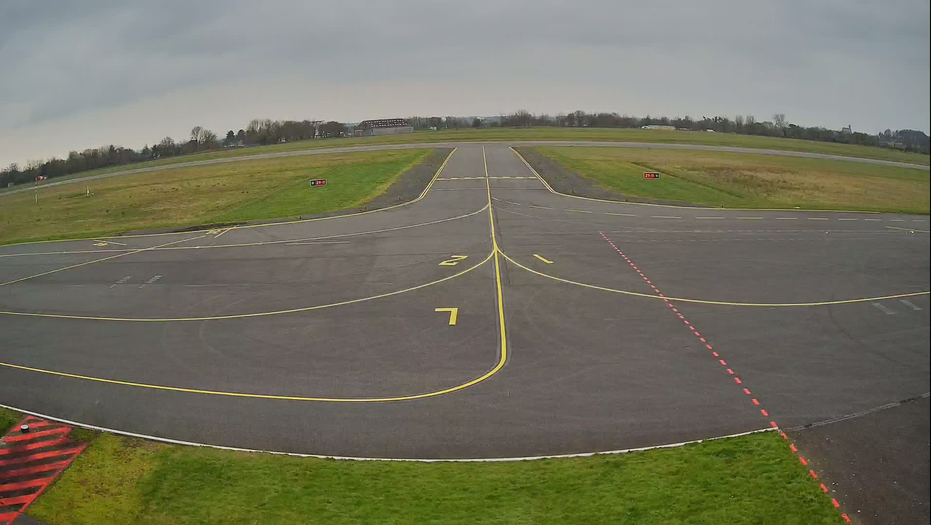 An overcast day at an airfield shows a paved area with yellow and red markings leading to a runway.