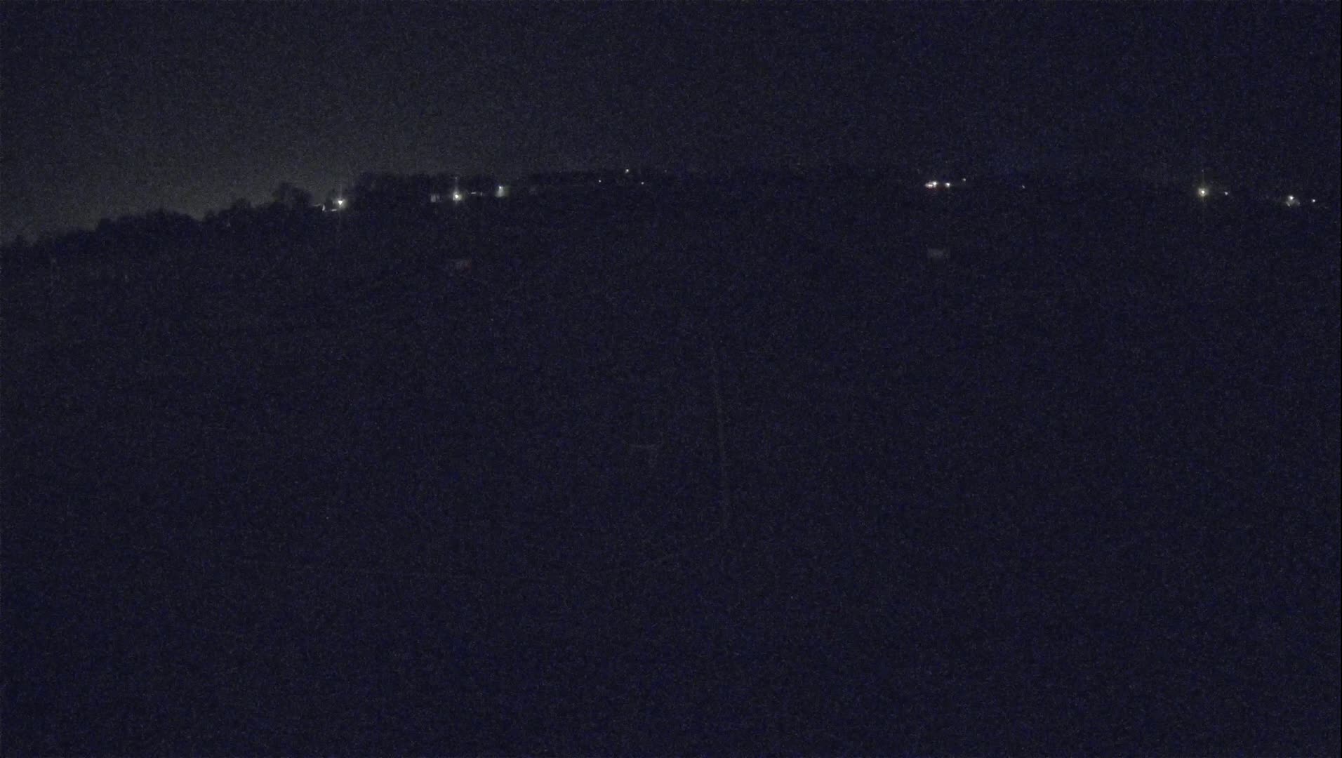 A dark, nighttime landscape shows a distant line of faintly lit buildings or structures across a mostly dark and indistinct horizon.