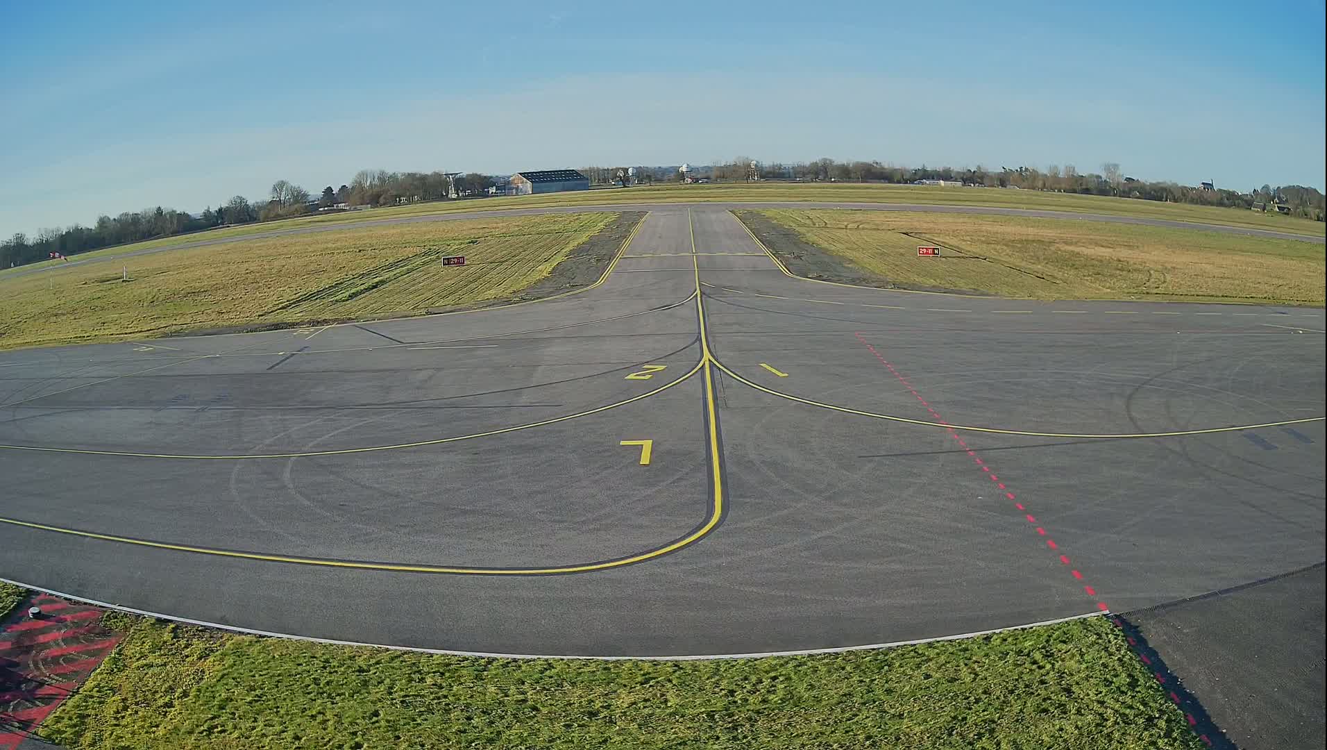 A mostly empty tarmac airport runway with short grass on either side, under a clear blue sky.