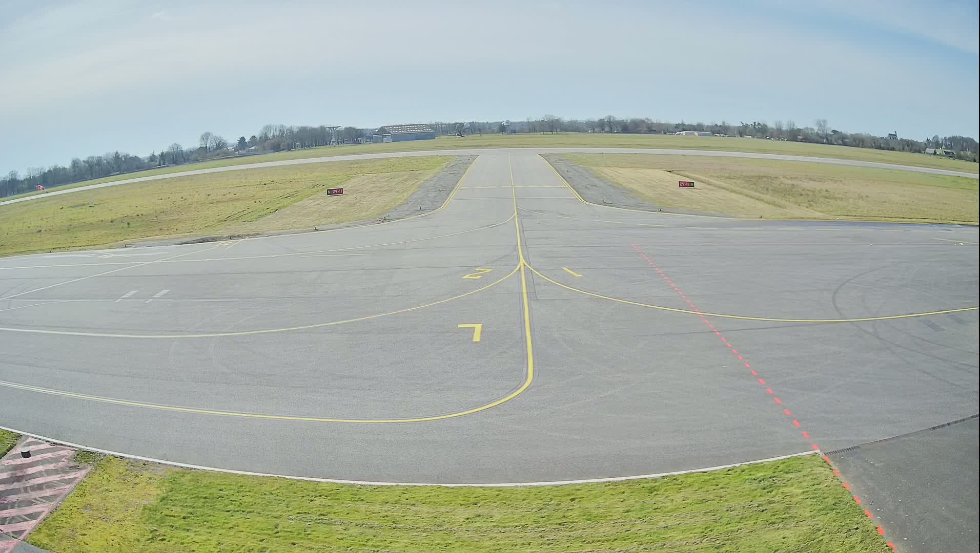 A paved airfield with yellow and red markings is shown under a partly cloudy sky.