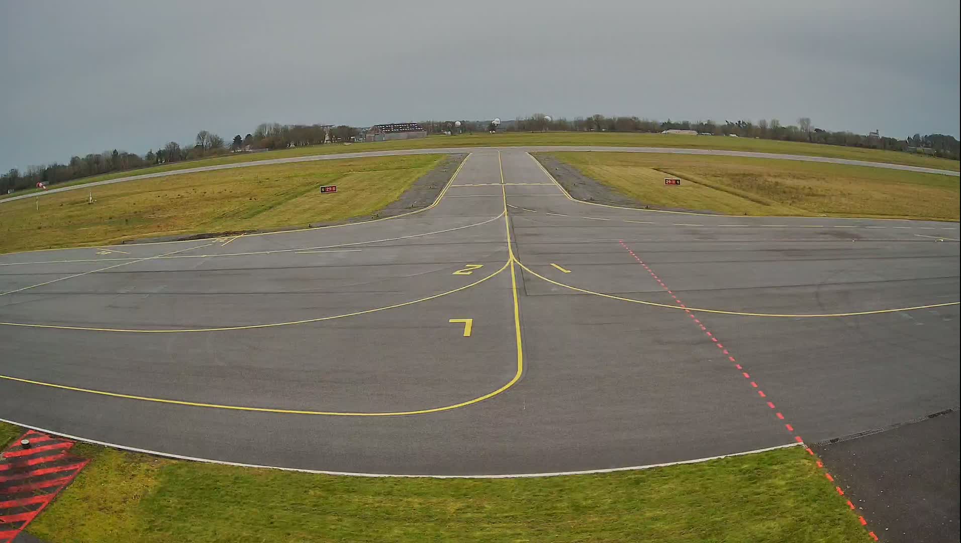 An airfield's tarmac, marked with yellow lines and numbers, extends toward a grassy runway under an overcast sky.