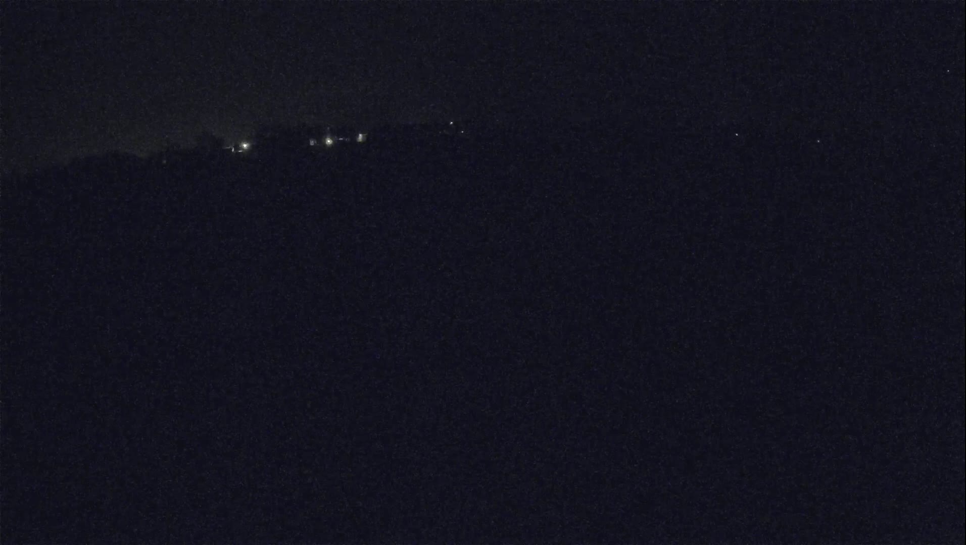 A nighttime, dark landscape shows a distant line of faint lights on a hill under a clear, dark sky.