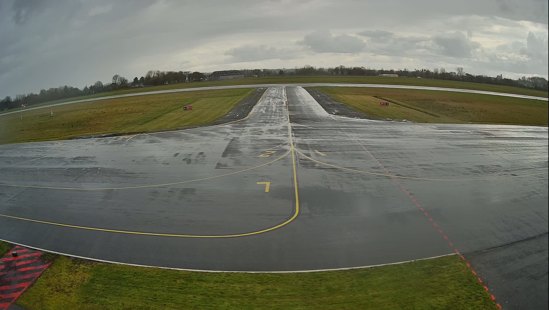 A wet, gray airport tarmac with yellow lines marking taxiways extends to a runway under an overcast sky.