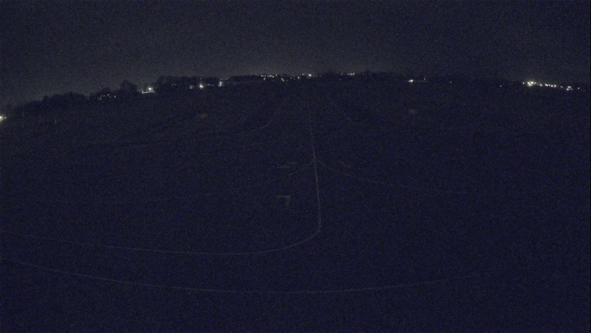 A nighttime, high-angle view shows a dark expanse of land with faint lights in the distance and indistinct lines suggesting roads or paths.