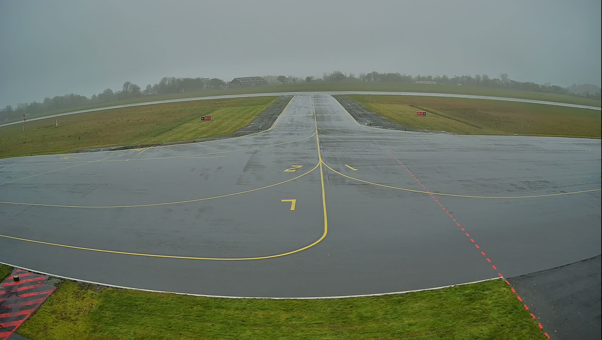 A wet airport runway with yellow and red markings is visible under an overcast sky.
