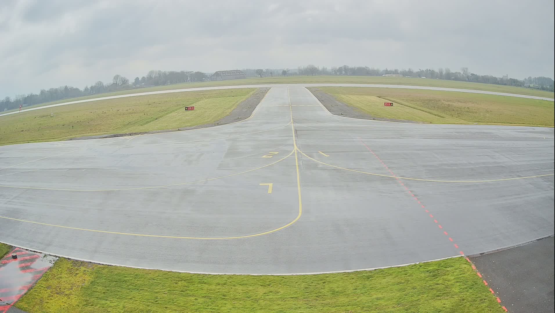 A wet airport runway with yellow lines marking taxiways and numbers indicating runway directions is shown under an overcast sky.