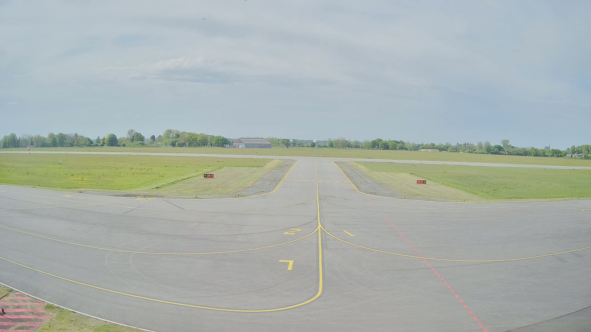 A paved airport runway and taxiway are visible under a partly cloudy sky, surrounded by grassy fields and trees in the distance.