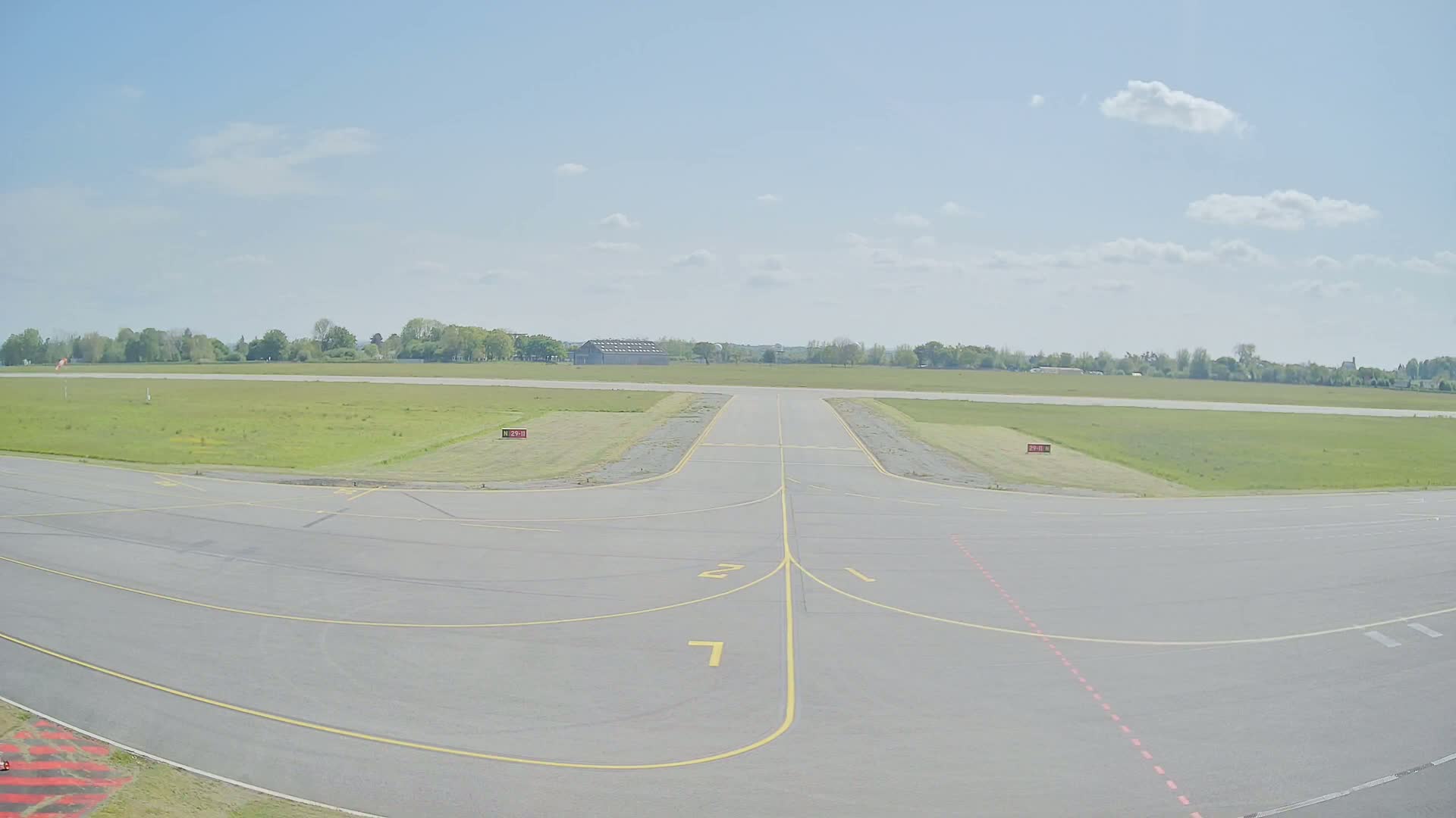 A tarmac runway with yellow lines, leading to a grassy airfield under a partly cloudy sky.