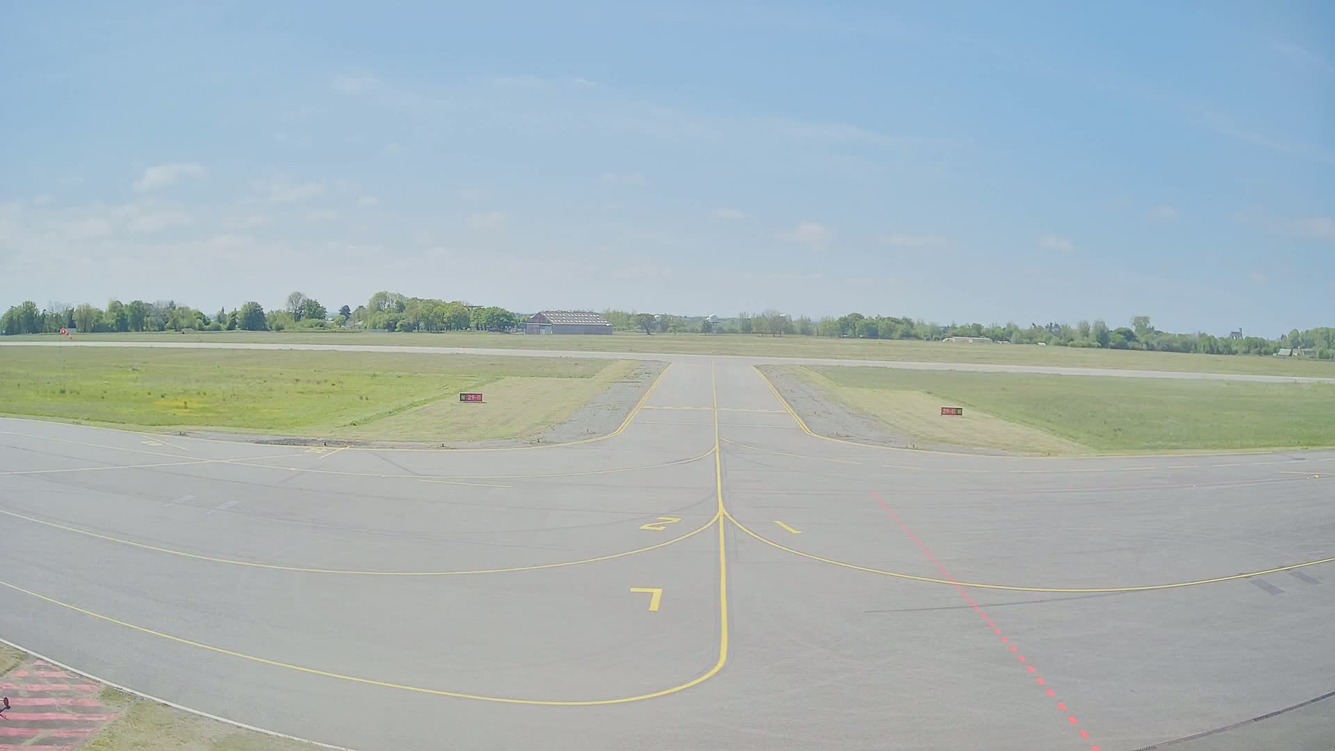 A paved runway at an airfield on a partly sunny day extends to a grassy field lined with trees.