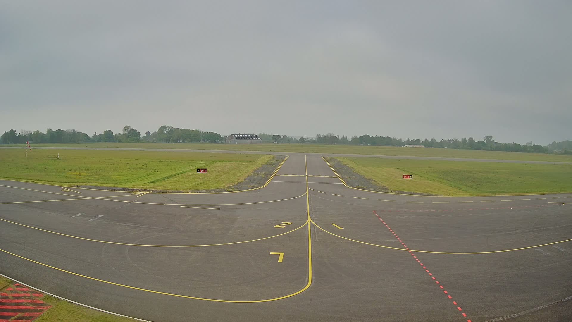 A tarmac runway at an airport on an overcast day, flanked by grassy fields and a line of trees in the distance.